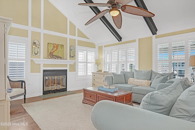 living room featuring ceiling fan, high vaulted ceiling, wood finished floors, wainscoting, and a glass covered fireplace