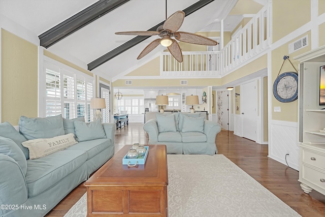 living area featuring a wainscoted wall, dark wood finished floors, visible vents, a ceiling fan, and beamed ceiling