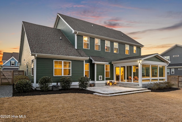 back of house at dusk with fence, a patio area, a sunroom, and roof with shingles