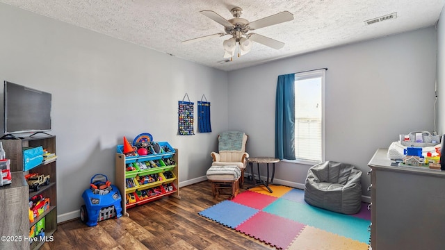 recreation room with a textured ceiling, ceiling fan, dark wood-type flooring, visible vents, and baseboards