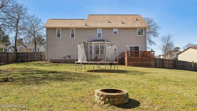 rear view of property with a deck, an outdoor fire pit, a fenced backyard, a lawn, and a trampoline