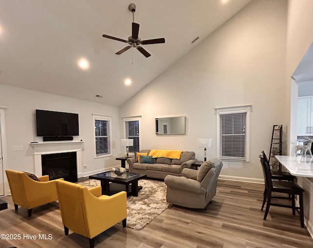 living area with ceiling fan, high vaulted ceiling, a fireplace, wood finished floors, and baseboards