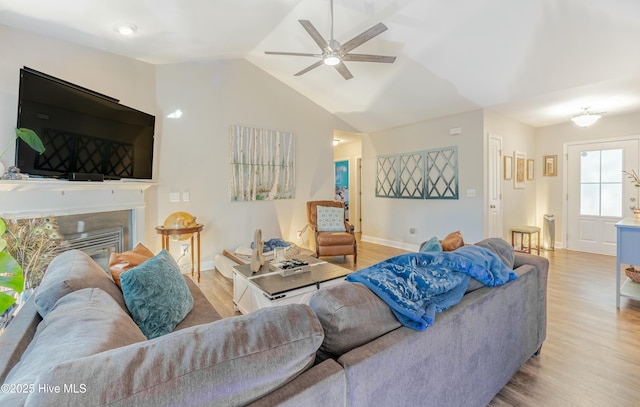 living area with light wood-style floors, a glass covered fireplace, vaulted ceiling, and a ceiling fan