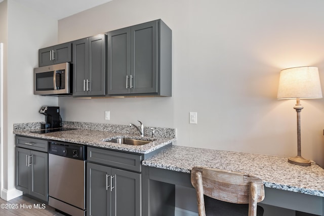 kitchen with light stone counters, gray cabinetry, a sink, appliances with stainless steel finishes, and built in desk