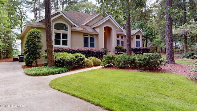 ranch-style house with a front lawn, a chimney, roof with shingles, concrete driveway, and stucco siding