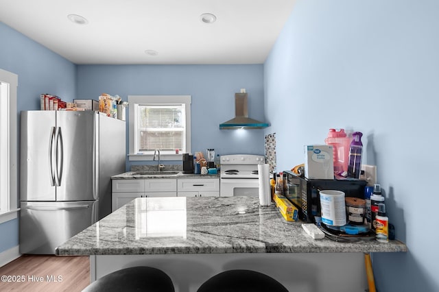 kitchen with white range with electric cooktop, a sink, freestanding refrigerator, wall chimney exhaust hood, and light stone countertops