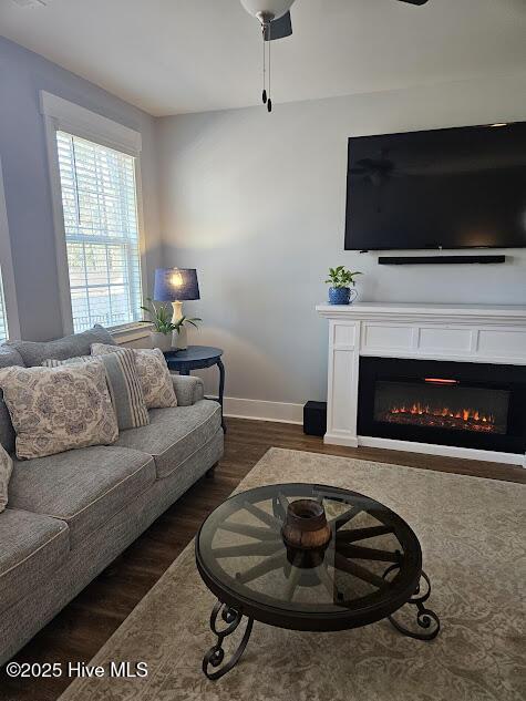 living room featuring a ceiling fan, a warm lit fireplace, dark wood finished floors, and baseboards