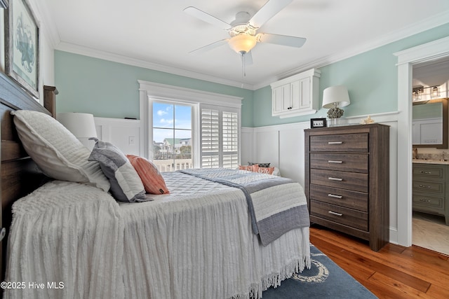 bedroom with a wainscoted wall, ensuite bathroom, wood finished floors, crown molding, and ceiling fan