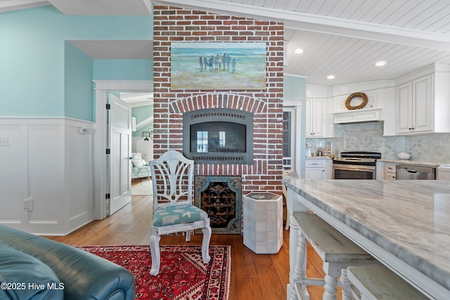 living area with a wainscoted wall, a brick fireplace, vaulted ceiling with beams, and light wood finished floors