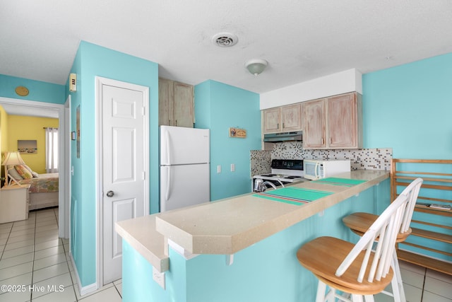 kitchen featuring under cabinet range hood, a peninsula, white appliances, light brown cabinetry, and a kitchen bar