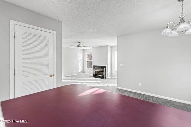 unfurnished living room featuring a textured ceiling, ceiling fan with notable chandelier, a fireplace with flush hearth, carpet flooring, and baseboards