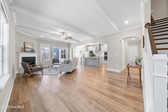 living room featuring arched walkways, crown molding, light wood-style flooring, a glass covered fireplace, and stairs