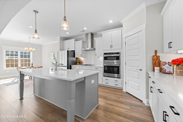 kitchen with light wood finished floors, appliances with stainless steel finishes, ornamental molding, wall chimney range hood, and backsplash