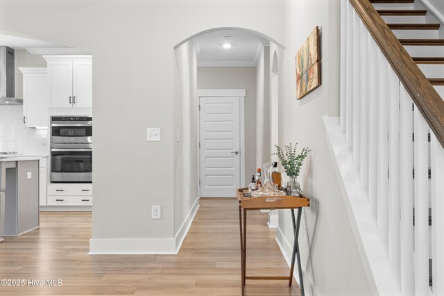 hall with arched walkways, baseboards, stairway, light wood finished floors, and crown molding