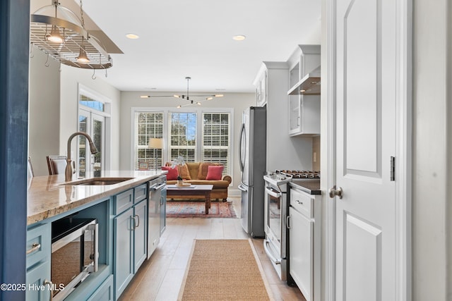 kitchen with blue cabinets, light wood-style flooring, a sink, appliances with stainless steel finishes, and wall chimney range hood