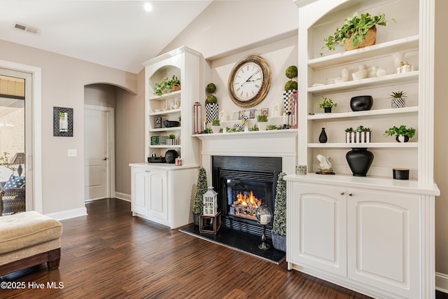 living area featuring built in features, dark wood finished floors, visible vents, a glass covered fireplace, and vaulted ceiling