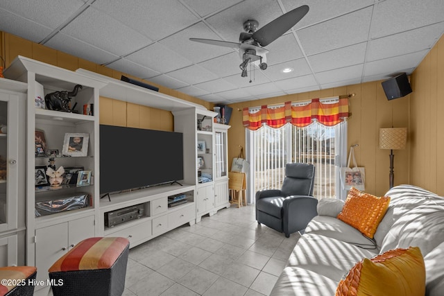 living area with ceiling fan, light tile patterned floors, wood walls, and a drop ceiling