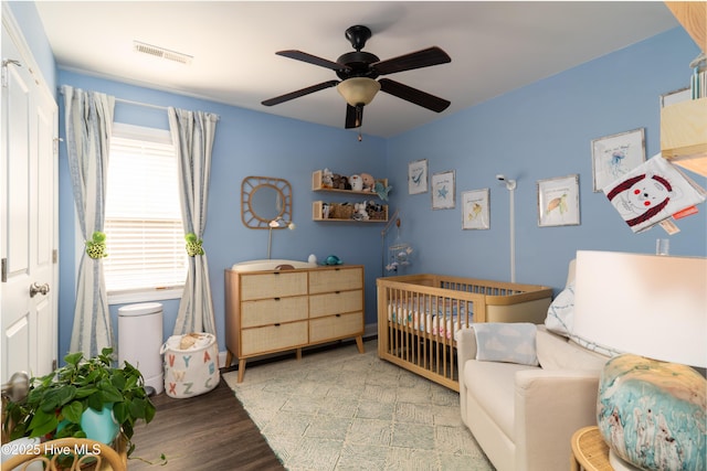 bedroom with ceiling fan, a nursery area, wood finished floors, and visible vents