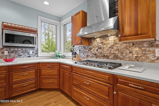 kitchen with brown cabinets, appliances with stainless steel finishes, light countertops, wall chimney exhaust hood, and light wood-type flooring