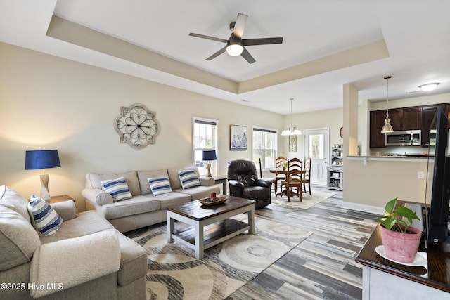 living area with a raised ceiling, light wood finished floors, ceiling fan with notable chandelier, and a wealth of natural light