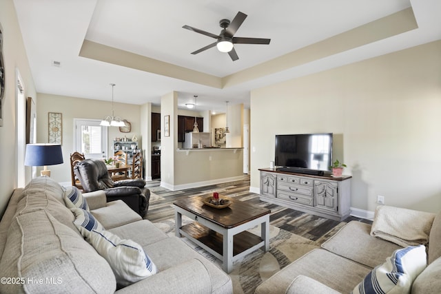 living room featuring dark wood finished floors, ceiling fan with notable chandelier, a raised ceiling, and baseboards