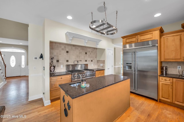 kitchen featuring a kitchen island with sink, light wood-style flooring, dark stone countertops, premium appliances, and tasteful backsplash