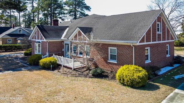 tudor home with brick siding, a chimney, a shingled roof, a wooden deck, and a front lawn