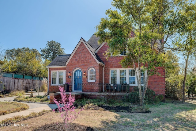 english style home featuring a front yard, fence, brick siding, and a shingled roof