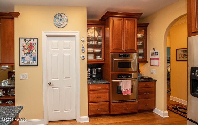 kitchen with arched walkways, stainless steel appliances, glass insert cabinets, light wood-style floors, and baseboards