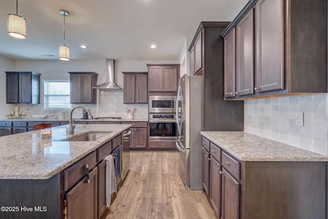 kitchen with a center island with sink, light wood finished floors, a sink, stainless steel appliances, and wall chimney range hood