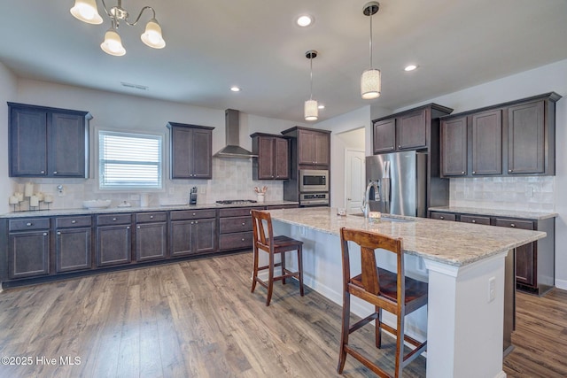 kitchen featuring wood finished floors, a sink, dark brown cabinets, appliances with stainless steel finishes, and wall chimney range hood