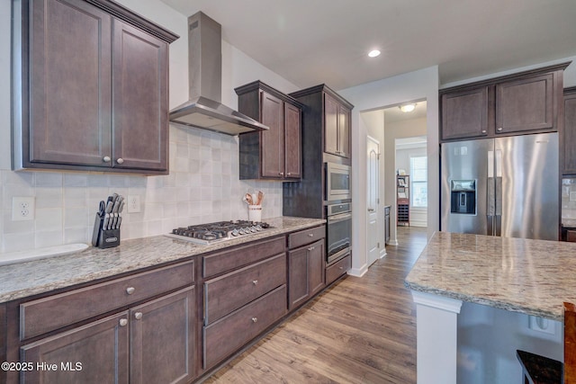 kitchen featuring wood finished floors, dark brown cabinetry, appliances with stainless steel finishes, wall chimney exhaust hood, and tasteful backsplash