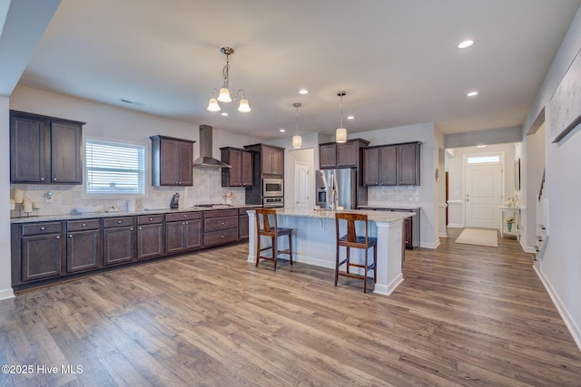 kitchen with wall chimney range hood, dark brown cabinetry, a kitchen breakfast bar, wood finished floors, and stainless steel appliances