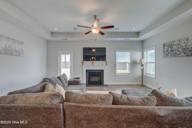 living area with a tray ceiling, a fireplace with flush hearth, wood finished floors, and baseboards