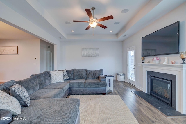 living room featuring a ceiling fan, a tray ceiling, a glass covered fireplace, wood finished floors, and recessed lighting