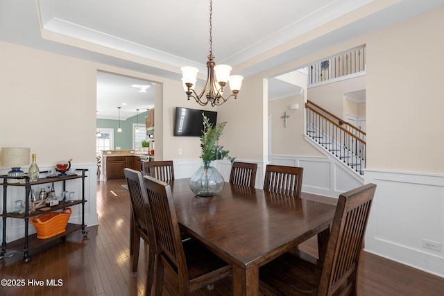 dining room with dark wood finished floors, a wainscoted wall, an inviting chandelier, stairs, and a decorative wall