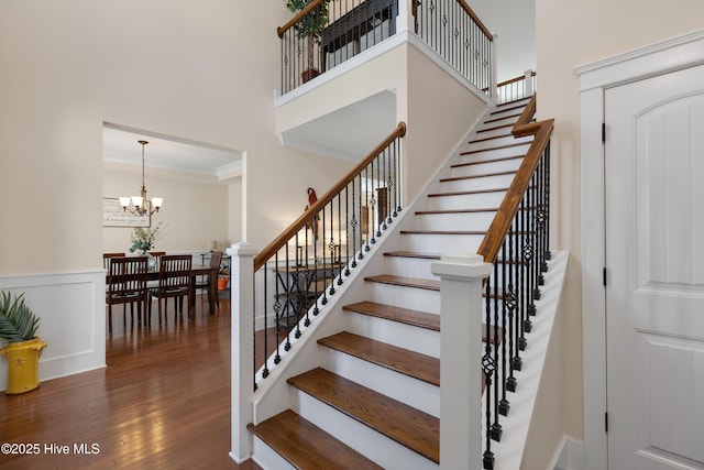 staircase featuring a wainscoted wall, ornamental molding, wood finished floors, an inviting chandelier, and a high ceiling