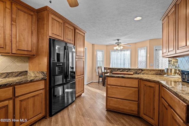 kitchen featuring a ceiling fan, black refrigerator with ice dispenser, light wood finished floors, brown cabinetry, and dark stone countertops