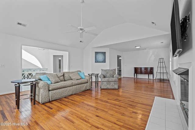 living room with light wood finished floors, visible vents, a glass covered fireplace, vaulted ceiling, and ceiling fan