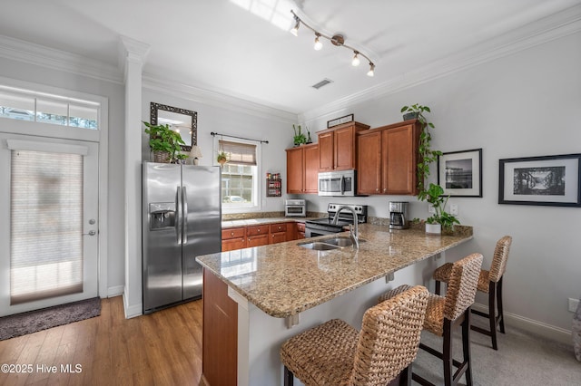 kitchen with brown cabinetry, appliances with stainless steel finishes, ornamental molding, a peninsula, and a sink