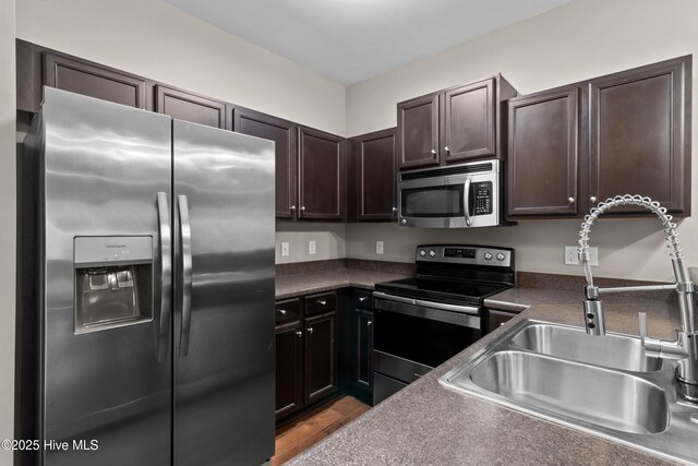 kitchen with dark countertops, wood finished floors, stainless steel appliances, dark brown cabinets, and a sink