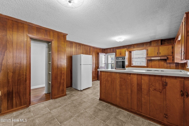 kitchen featuring brown cabinets, freestanding refrigerator, light countertops, under cabinet range hood, and black oven