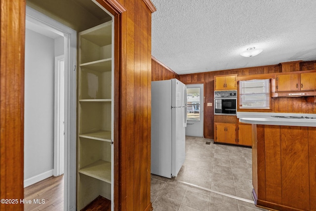 kitchen featuring oven, wooden walls, under cabinet range hood, freestanding refrigerator, and brown cabinetry