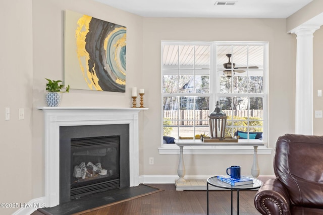 living area with wood finished floors, visible vents, baseboards, a glass covered fireplace, and decorative columns