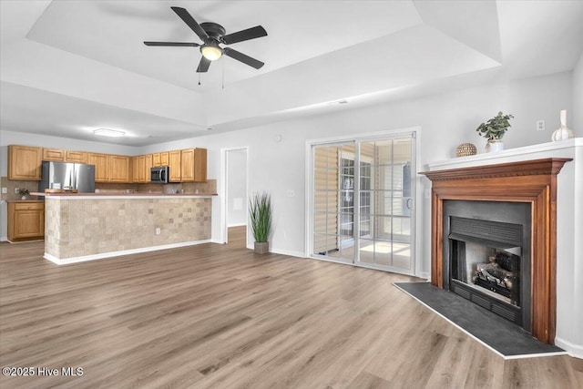 unfurnished living room featuring a fireplace with raised hearth, a ceiling fan, baseboards, a tray ceiling, and light wood finished floors