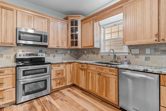 kitchen featuring appliances with stainless steel finishes, glass insert cabinets, light brown cabinets, a sink, and light wood-type flooring