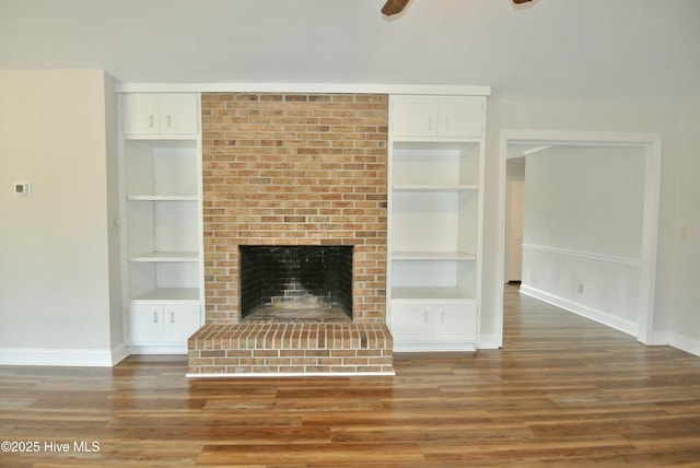 unfurnished living room with baseboards, ceiling fan, wood finished floors, a brick fireplace, and built in shelves