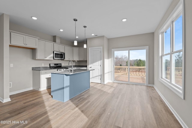 kitchen featuring light wood finished floors, stainless steel appliances, hanging light fixtures, a kitchen island with sink, and baseboards