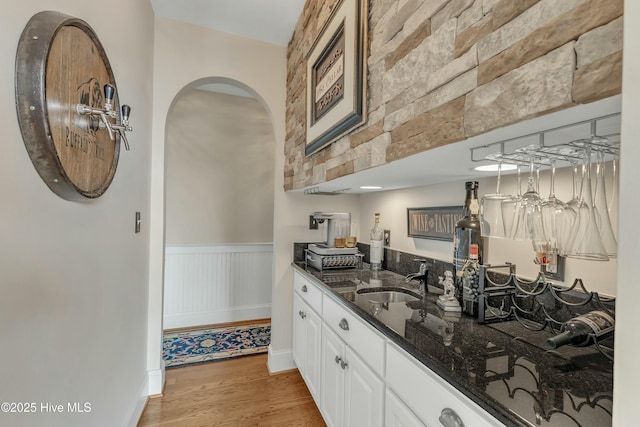 kitchen with a sink, white cabinetry, light wood-type flooring, wainscoting, and dark stone countertops