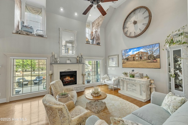 living area with a warm lit fireplace, a ceiling fan, wood finished floors, a high ceiling, and recessed lighting